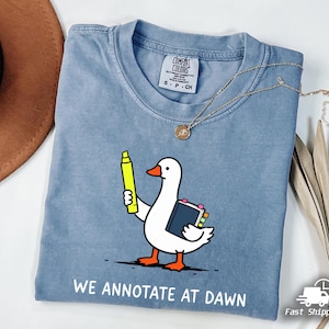 May include: A blue short-sleeved t-shirt featuring a graphic of a white goose holding a yellow highlighter and a book with colorful tabs. Below the graphic, the text reads "WE ANNOTATE AT DAWN" in white capital letters. The shirt has a crew neckline.