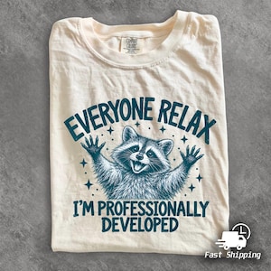 Puede incluir: Una camiseta color crema con un gráfico en verde azulado oscuro de un mapache con los brazos levantados. El texto "EVERYONE RELAX I'M PROFESSIONALLY DEVELOPED" aparece en letras verde azulado oscuro, rodeado de pequeños iconos de estrellas.
