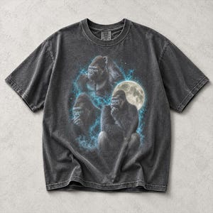 May include: A charcoal grey short-sleeve t-shirt featuring a graphic print of three gorillas in various poses. The design includes a bright full moon and glowing blue lightning-like energy surrounding the gorillas, set against a dark background.