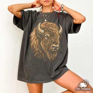 May include: A charcoal gray short-sleeve t-shirt featuring a large, detailed graphic print of a brown bison head on the front. The bison is depicted in profile with textured fur and curved horns. The shirt has a crew neckline and a relaxed fit.