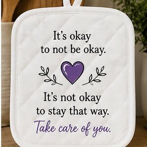 Könnte beinhalten: Weißer, gesteppter Topflappen mit Aufhängeschlaufe. Der Text lautet: "It's okay to not be okay. It's not okay to stay that way. Take care of you." Mit einem lila Herz und Blättern.