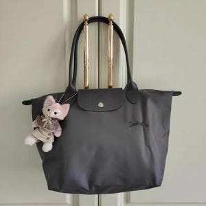May include: A gray tote bag with black leather handles and a flap closure. A small plush toy in a pink and brown outfit hangs from the bag. The bag is hanging on a gold-colored rod.
