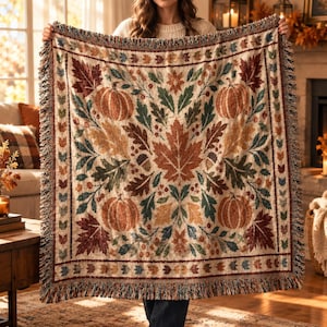 May include: A woven throw blanket featuring a symmetrical autumn pattern with orange pumpkins, brown maple leaves, green foliage, and small acorns on a cream background. The blanket has a decorative border of small leaves and a fringed edge.