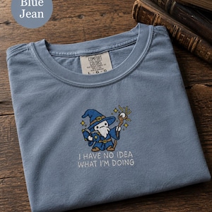 May include: A blue jean coloured t-shirt featuring an embroidered graphic of a wizard holding a staff and a book. The text "I HAVE NO IDEA WHAT I'M DOING" is embroidered in white below the wizard. The shirt has a Comfort Colors brand label.