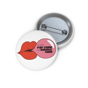 May include: A round white pin-back button featuring a graphic of red lips blowing a pink bubble gum bubble. Inside the bubble, the black text reads: raised on POP PUNK and BUBBLE GUM. The back of the button shows a silver metal pin mechanism.