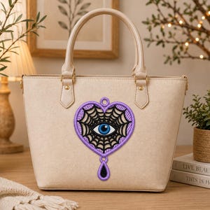 Op de afbeelding: Een beige gestructureerde handtas met een centraal geborduurd embleem in de vorm van een hart met een zwart spinnenwebpatroon en een blauw oog in het midden. Het embleem is omlijnd met lichtpaarse draad en heeft een kleine zwarte druppel.