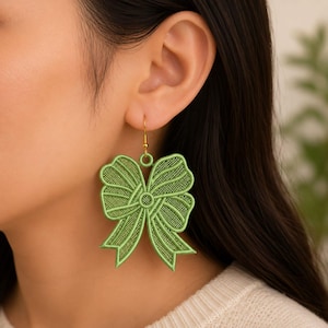 Può includere: Un orecchino a forma di fiocco ricamato verde chiaro con una texture dettagliata simile a una rete e un gancio in metallo dorato. Il design del fiocco presenta bordi cuciti definiti e due nastri pendenti, creando un aspetto strutturato.