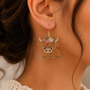 May include: A gold-toned dangle earring featuring a brown Highland cow with a textured, lace-like design. The cow has a floral crown with pink, white, and green flowers on its head and is suspended from a simple gold-colored metal hook.