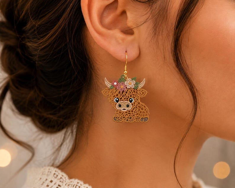 May include: A gold-toned dangle earring featuring a brown Highland cow with a textured, lace-like design. The cow has a floral crown with pink, white, and green flowers on its head and is suspended from a simple gold-colored metal hook.