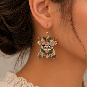 May include: A dangling earring featuring a tan, lace-like embroidered highland cow design. The cow has brown horns, black and white eyes, and is adorned with green holly leaves and small red berries around its head and neck on a gold-tone hook.