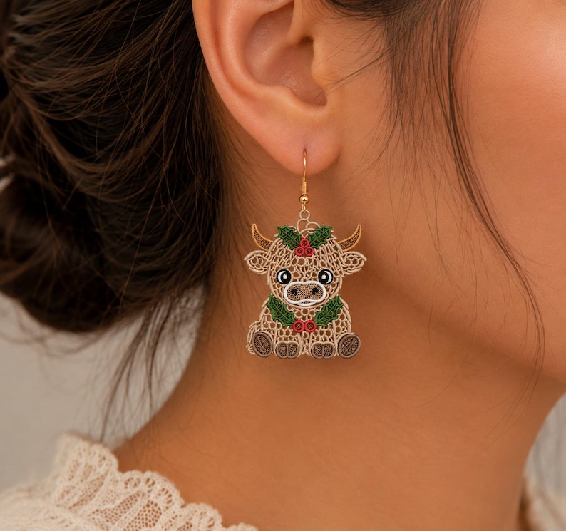 May include: A dangling earring featuring a tan, lace-like embroidered highland cow design. The cow has brown horns, black and white eyes, and is adorned with green holly leaves and small red berries around its head and neck on a gold-tone hook.