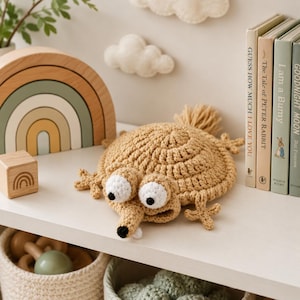 May include: A tan crochet creature with a round body, a pointed snout, a small white tooth, and two large white eyes with black pupils. The creature has four splayed limbs and a tuft of yarn at the back, crafted from textured yarn.