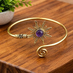 May include: A gold-toned metal cuff bracelet featuring a central sunburst motif with a round, purple gemstone at its center. The bracelet has a textured, open-ended design with one end terminating in a spiral coil and the other in the sunburst.