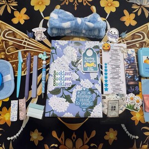 May include: A collection of stationery and self-care items including a floral notebook with stickers, a blue polka dot headband, a blue facial sponge, a pen, a grapefruit-scented hand sanitizer, a bookmark, and small individually wrapped orange tablets.