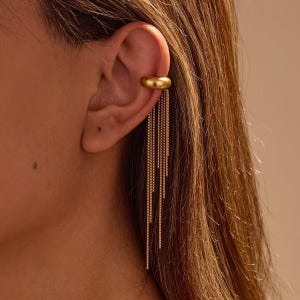 Op de afbeelding: Een goudkleurige ear cuff met een gladde, ronde band die aan het bovenste kraakbeen van het oor wordt bevestigd. Aan de band hangen meerdere fijne, bungelende kettingstrengen van verschillende lengtes die verticaal naar beneden vallen.
