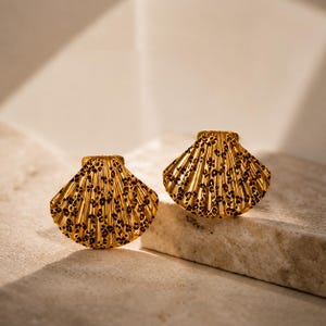 May include: Two gold-coloured seashell-shaped earrings with a textured surface and dark brown spots. The earrings are displayed on a light-coloured stone surface, highlighting their intricate design and metallic sheen.