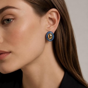 May include: A circular stud earring featuring a polished gold-tone center dome surrounded by a smooth, glossy blue outer ring. The earring is secured to the earlobe, showcasing a contrast between the metallic gold finish and the deep blue border.