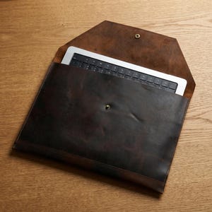 May include: A dark brown leather envelope-style sleeve containing a silver laptop with a black keyboard. The sleeve features a pointed flap with a single brass-colored metal snap closure and a matching snap on the front body for secure fastening.