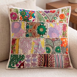 May include: A square throw pillow featuring a patchwork design of embroidered fabric panels. The surface displays a variety of colorful floral, paisley, and geometric patterns in shades of orange, green, purple, pink, and gold with white piping.