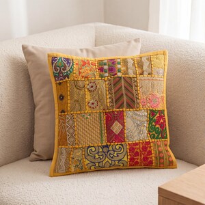May include: A square decorative throw cushion featuring a patchwork design of various embroidered fabrics in shades of gold, yellow, red, green, and purple. The front panel displays intricate patterns, metallic thread accents, and beadwork.