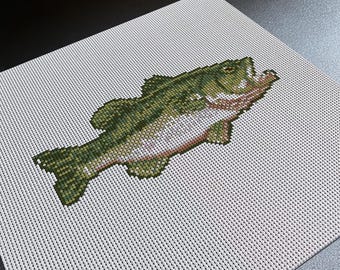 Puede incluir: Un bordado de punto de cruz sobre tela Aida blanca con un diseño detallado de un pez. El pez está representado en tonos verde oscuro, oliva y blanco, con una boca abierta visible y escamas texturizadas creadas por hilos de punto de cruz.