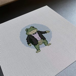 May include: A square piece of white needlepoint canvas featuring a cross-stitch design of a green frog wearing a black tuxedo jacket and a light purple shirt. The frog is centered against a circular light blue background on the fabric mesh.