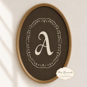 May include: An oval-shaped wall art piece featuring a dark brown background with a cream-coloured letter A in the centre, enclosed by a decorative white filigree border. The artwork is encased in a thin, metallic gold frame with a textured edge.