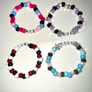 May include: Four circular bracelets made of colorful plastic pony beads. One bracelet features pink, blue, and black beads with the name MIZI. Another has purple, black, and clear star beads with SUA. A third has red, white, and black beads with IVAN. The last has blue, teal, and clear star beads with TILL.