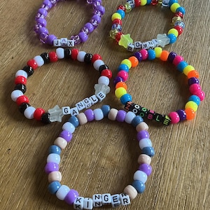 May include: Five elastic bracelets made of colorful plastic pony beads. Each bracelet features white letter beads spelling JAX, POMNI, GANGLE, ZOOBLE, or KINGER. Some bracelets include star-shaped beads, while others use solid or translucent round beads.