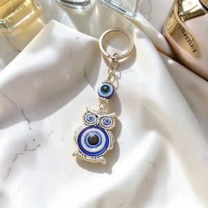 Puede incluir: Llavero de búho plateado con diseño de ojo turco azul y blanco. El búho tiene ojos grandes y redondos y un patrón de plumas detallado. Una cuenta de ojo turco más pequeña cuelga encima del búho.