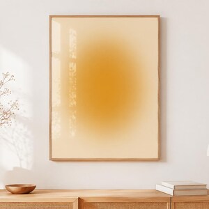 May include: A framed art print featuring a warm, gradient design in shades of yellow and orange. The artwork is displayed on a white wall, with a wooden frame. The room is decorated with a vase of dried flowers, a wooden bowl, and a mushroom-shaped lamp.