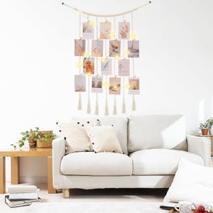 May include: A macrame photo display with hanging picture frames and fairy lights. The wall decor features a natural, off-white rope design with wooden beads and tassels. The frames hold various photos, creating a bohemian aesthetic.