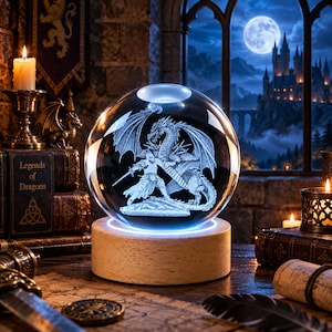 May include: A clear glass sphere featuring a laser-etched 3D engraving of a knight fighting a dragon, mounted on a circular light-up wooden base. The sphere displays a detailed scene of a warrior with a sword facing a winged creature.