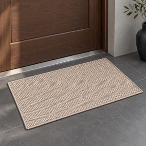 May include: A rectangular doormat featuring a woven beige and white herringbone pattern. The mat has a finished bound edge and a textured surface, providing a neutral color palette that complements the surrounding dark wood door and gray tile floor.