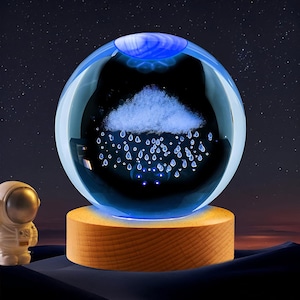 May include: A spherical glass lamp featuring an internal 3D laser-etched design of a cloud with falling raindrops. The sphere is illuminated with a blue light and rests on a cylindrical light-colored wooden base, accompanied by a small astronaut figure.