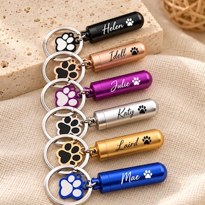 May include: A set of six cylindrical metal cremation urn keychains in black, rose gold, purple, silver, gold, and blue. Each keychain features a paw print charm and is engraved with a name: Helen, Idell, Julie, Katy, Laird, and Mae.