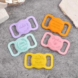 May include: Five silicone pet collar tags in lavender, orange, mint green, pink, and yellow. Each tag features a central circular area with custom engravings including names like Violet, Abby, Sophia, and Jacob, along with phone numbers and addresses.