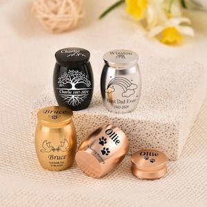May include: Four small metal cremation urns in black, silver, gold, and rose gold finishes. The black urn features a tree design and the name Charlie. The silver urn shows a rainbow and the text Best Dad Ever. The gold urn displays doves and the name Bruce. The rose gold urn is engraved with the name Ollie and paw prints.