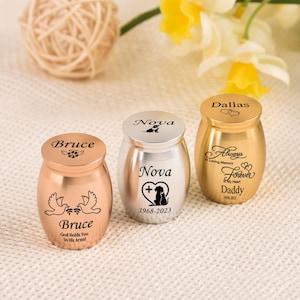 Può includere: Tre piccole urne cinerarie in metallo con finiture in oro rosa, argento e oro. Ogni urna presenta incisioni personalizzate che includono nomi come Bruce, Nova e Dallas, insieme a grafiche commemorative come colombe, la sagoma di un cane e cuori.
