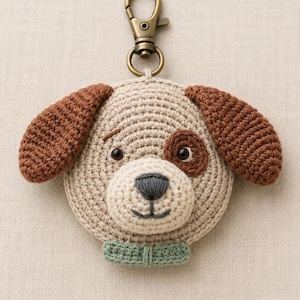 Puede incluir: Un llavero de ganchillo con forma de cabeza de perro que presenta una cara beige, orejas marrones, un parche marrón en el ojo, ojos de plástico negro, nariz y boca bordadas en gris oscuro y un collar verde claro con un mosquetón de bronce.