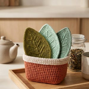 May include: A set of three crocheted leaf-shaped coasters in shades of olive green, mint green, and sage green, stored inside a rectangular crocheted basket with a terracotta-colored base and a cream-colored rim.