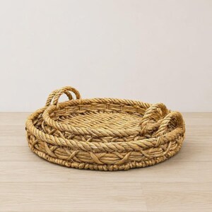 May include: A set of two round, woven trays made from natural light brown seagrass or similar fiber. Each tray features a thick, braided rope-style rim and two integrated handles on opposite sides, with a circular base and textured weave.