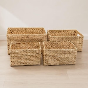 May include: A set of four rectangular storage baskets woven from natural light brown dried plant fibers. The baskets feature a consistent braided pattern throughout the sides and base, with integrated cutout handles on the shorter ends.