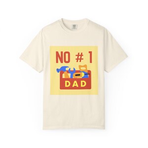 May include: A cream-colored short-sleeve t-shirt featuring a yellow square graphic on the chest. The graphic displays the red text NO # 1 above a red toolbox containing blue and yellow tools, with the yellow text DAD printed on the toolbox front.