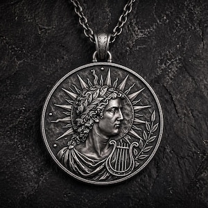 Puede incluir: Un collar con colgante plateado que presenta un relieve detallado de una figura clásica con una corona de laurel y una lira. El colgante circular tiene un diseño de rayos de sol alrededor de la cabeza de la figura. La cadena también es plateada.