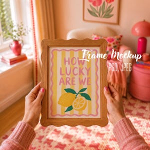 Può includere: Una stampa incorniciata in legno con la scritta "HOW LUCKY ARE WE" e un'illustrazione di limone. La stampa ha uno sfondo a righe gialle e bianche con bordi rosa. La cornice è tenuta da una persona che indossa un maglione rosa.