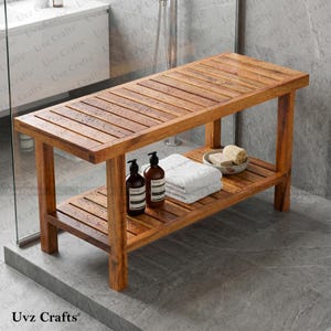 May include: A rectangular wooden shower bench with a slatted top and a lower shelf. The bench features a warm brown finish and is holding two dark brown pump bottles, a folded white towel, and a small bowl containing a bar of soap.