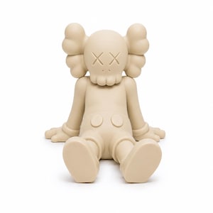 Op de afbeelding: Een beige vinyl figuur in een zittende positie met uitgestrekte armen en benen. De figuur heeft grote, afgeronde oren, doorgestreepte ogen en een schedelachtig gezicht. De figuur draagt een korte broek met twee ronde knopen.