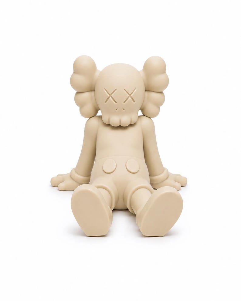 Op de afbeelding: Een beige vinyl figuur in een zittende positie met uitgestrekte armen en benen. De figuur heeft grote, afgeronde oren, doorgestreepte ogen en een schedelachtig gezicht. De figuur draagt een korte broek met twee ronde knopen.