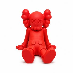 Op de afbeelding: Een effen rode vinyl figuur in een zittende positie. De figuur heeft grote, afgeronde oren, doorgestreepte ogen en een kleine mond. De figuur draagt een korte broek en heeft zijn handen op zijn benen. Een verzamelobject.